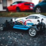 Close-up of a remote control car chassis with colorful wires on asphalt, with blurred cars in background.